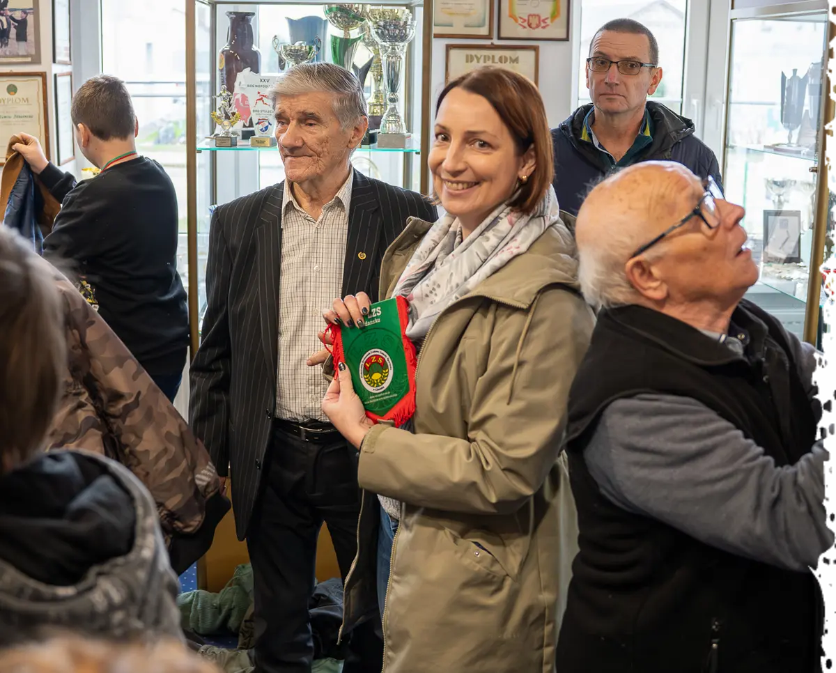 Pani z muzeum pokazuje zielony proporczyk LZS dwóm starszym panom na tle gablot z pucharami